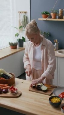 Orta yaşlı, beyaz bir kadının yeşil bitkiler ve doğal ışıkla süslenmiş, modern mutfağın ahşap tezgahında patlıcan keserken çekilmiş orta yaş fotoğrafı.