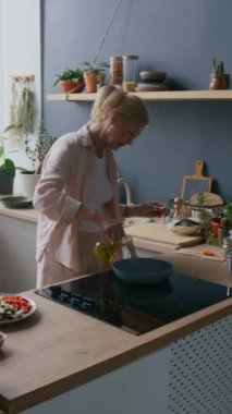 Orta yaşlı, rahat günlük giyimli, rahat bir kadın rahat mutfağın yanında duruyor ve tavaya zeytinyağı ekliyor. Gündüz sağlıklı yemek pişirirken, dikey çekim.