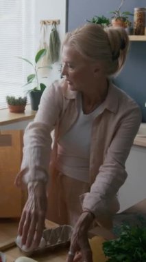 Yetişkin, beyaz bir kadının güneş ışığı altındaki mutfakta bitkilerle, ahşap tezgahlarla ve iyi düzenlenmiş yemek pişirme gereksinimleriyle kahverengi kağıt poşetlerden taze ürünler çıkardığı orta boy bir fotoğraf.