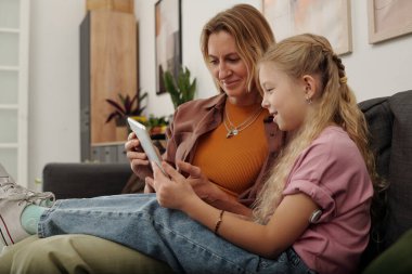 Annesiyle birlikte dijital tablet kullanan mutlu genç kızın yan görünüm portresi rahat evdeki kanepede oturup internetteki fotokopi alanlarını karıştırıyor.