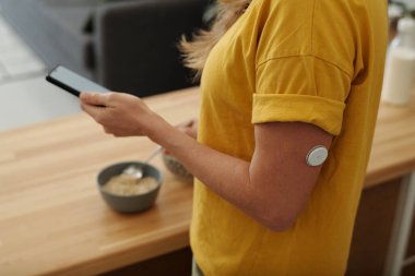 Diabet hastası yetişkin bir kadının sabah sağlıklı kahvaltı hazırlarken kol fotokopi uzayı üzerindeki CGM sensörüne odaklanarak akıllı telefon kullanmasını yakından çek. 