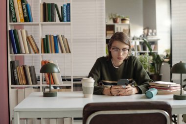 Genç bir kızın kız öğrenci portresi. Üniversite kütüphanesinde telefon kullanıyor. Pembe aksanlı en az ayarda.