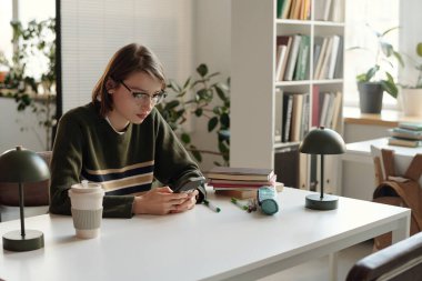 Sınıfta ya da öğrenci salonunda otururken telefon kullanan ve gözlük takan genç bir kadının portresi 