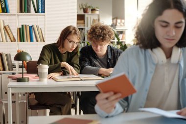 Üniversite kütüphanesinde kitap üzerinde çalışan iki öğrencinin ön plandaki fotokopi alanında oturan genç çocukla sınavlara hazırlanırken omuz üstünden çekilen fotoğrafı. 