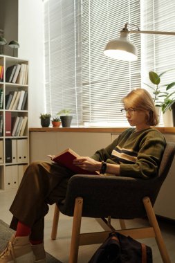 Genç bir kadının kitap okuduğu dikey yan manzara portresi. Sıcak, rahat bir koltukta oturmuş, sessiz bir hobiden keyif alıyor.