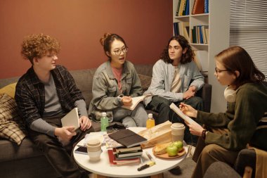 Yüksek açılı bir bakış açısı, birlikte ders çalışan bir grup gencin, rahat kütüphane salonunda akşam vakti, düşük ışıkla birlikte yuvarlak masa başında oturmalarını sağlıyor.