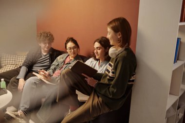 Bir grup öğrenci kanepede birlikte ders çalışıyor ve rahat üniversite salonunda düşük ışıkla ödev yapıyorlar.