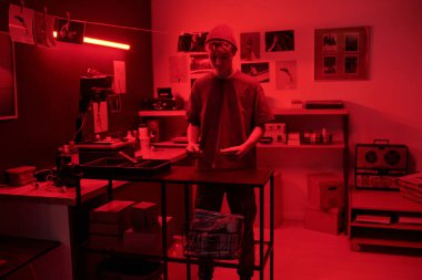 Karanlık odada çalışan genç bir adamın tam boy portresi kırmızı ışıkla aydınlatılıyor ve yaratıcı stüdyo fotokopi alanında analog fotoğraflar geliştiriyor.