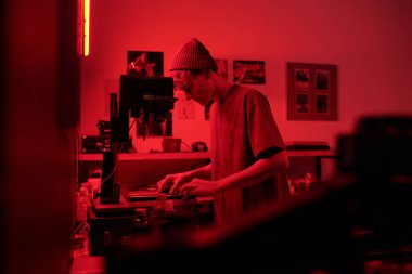 Karanlık odada ekipmanla çalışan genç bir çocuğun karamsar yan görünüm portresi stüdyo fotokopisinde kırmızı ışıkta genç fotoğrafçıların fotoğraflarını basıyor.