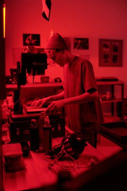 Geniş ekipmanlarla çalışan genç fotoğrafçının dikey yan görünüm portresi. Stüdyoda kırmızı ışık genç fotoğrafçı tarafından aydınlatılan karanlık odada fotoğraf baskıları geliştirirken.