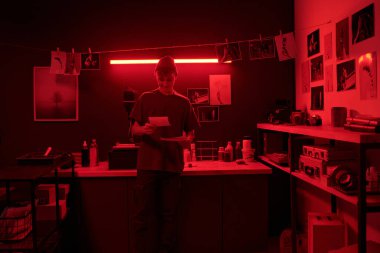 Elinde fotoğraflarla dolaşan gülümseyen bir gencin ön manzara portresi stüdyo fotokopi odasında kırmızı ışıkta genç bir fotoğrafçı tarafından aydınlatılırken karanlık odada fotoğraf çektiriyordu.