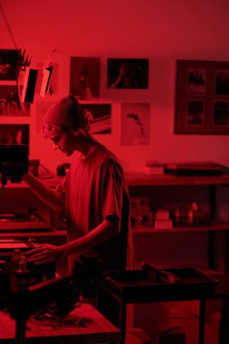 Gencin dikey portresi, daha büyük ekipmanları ayarlarken yaratıcı stüdyodaki kırmızı ışık genç fotoğrafçısının karanlık odada fotoğraf geliştirme üzerinde çalışması.