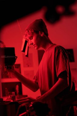 Kırmızı ışık yaratıcı stüdyo ortamının aydınlattığı karanlık odada parmak izlerini geliştirirken genç fotoğrafçının karamsar yakın plan portresi büyültülmüş.