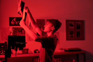 Genç fotoğrafçının karanlık oda fotokopi odasında kırmızı ışıkta kurutmak için yeni geliştirilmiş fotoğraf baskısını asmasının yaratıcı bir yan görüntüsü.