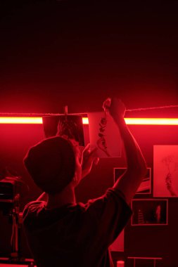 Huysuz arka plan görüntüsü, genç bir adamın, yaratıcı stüdyoda çalışan genç bir fotoğrafçının kırmızı ışıkta kurutma çizgisinde asılı duran fotoğraflara uzanmasını çekti.