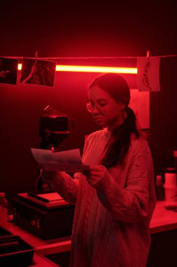 Analog fotoğraf baskılarıyla çalışan ve kırmızı ışık yaratıcı stüdyo ortamının aydınlattığı karanlık odada resim seçen genç bir kızın dikey portresi.