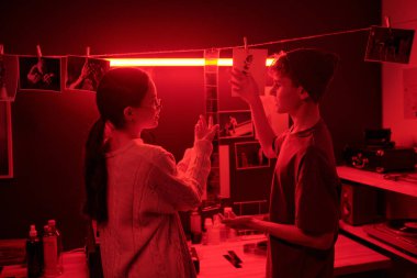 Artistic portrait of teenage boy and girl discussing developed film strip hanging over red neon light in darkroom young photographers together in creative studio clipart