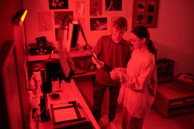 Karanlık odada analog fotoğraf baskıları geliştirirken aynı zamanda gülümseyen oğlan ve kızın film şeritlerine baktığı yüksek açı görüntüsü