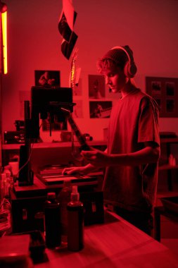 Kulaklık takan genç bir çocuğun dikey yan görüntüsü. Elinde gelişmiş film şeridi tutarken, karanlık odada kırmızı neon ışıkla büyüyen bir fotoğrafla çalışıyor.