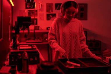 Kırmızı ışıkla aydınlatılan karanlık odada çalışırken ve kulaklıkla müzik dinlerken kimyasal tepsiler kullanarak fotoğraf baskıları geliştiren genç bir kızın portresi.