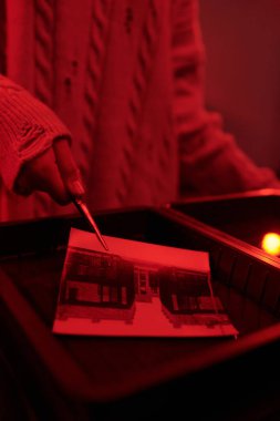 Kırmızı ışık fotokopi alanı tarafından aydınlatılan karanlık odada siyah beyaz fotoğraf baskısı geliştirmek için cımbız kullanan kızın yakınlaşması