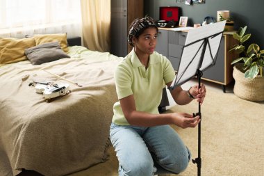 Genç insan rahat yatak odasında müzik sehpası, rahat atmosfer, özel ışıklandırma, kişisel eşyalar ve davetkar çevre ile gitar çalışıyor.