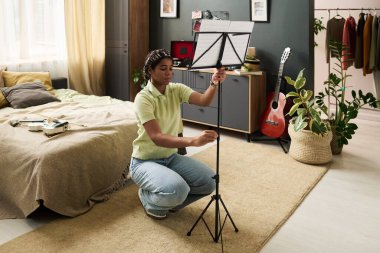 Rahat giyimli kız rahat bir yatak odasında gitar ve dekor ekleyen bitkilerle müzik standı kuruyor. İşine odaklanmış görünüyor.