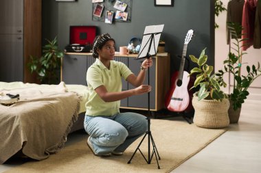 Kız rahat yatak odasında müzik standını odaklı ifade, gitar ve bitki süsleme odası ile ayarlıyor. Bağlılık ve hazırlık hissi sahneden yayılıyor.