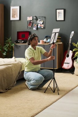 Genç Afro-Amerikalı kız, elinde gitar, pikap ve dekor unsurları ile donatılmış rahat yatak odasında müzik kağıdını tutarken yerde diz çöküyor.