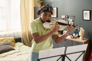 Genç Afro-Amerikalı kız rahat bir odada keman çalıyor, iyi ışıklandırılmış müzik standı ve çeşitli dekorlarla. Yumuşak doğal ışıklandırma, müzik çalışmalarının sıcak atmosferini arttırır.