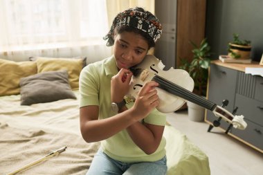 Genç kız yatak odasında keman çalıyor düşünceli bir ifadeyle, rahat dekor ve bitkilerle çevrili, müziğe olan aşkını gösteriyor.