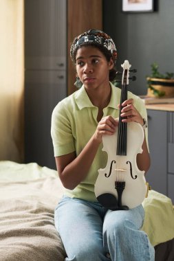 Genç kız yatakta oturup kemanını tutarken özenle arka planda bitkiler ve ahşap elementlerle dolu rahat bir odada yansıtıyor.