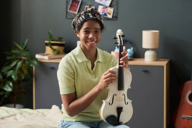 Gülümseyen kız rahat yatak odasında kemanını tutarken nazikçe, arka planda görünen çeşitli kişiselleştirilmiş dekorasyonlar, sıcak bir atmosfer yaratıyor.