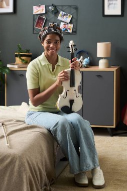 Genç kız elinde keman tutarken yatakta oturuyor ve gülümsüyor. Konforlu oturma odası arka planında saksı bitkileri, duvarda resimler ve sıcak atmosfer yaratan mobilyalar.