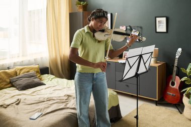 Gençler rahat oturma odasında arka planda modern dekor ve gitarla keman çalarken. Odaklanmış ifade, sıcak atmosfer