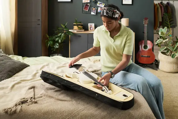 Kız yatakta oturup gitar ve bitkilerle rahat yatak odasında ukulele akort ediyor. Duvar dekorasyonu ve doğal elementler atmosfere sıcaklık katar.