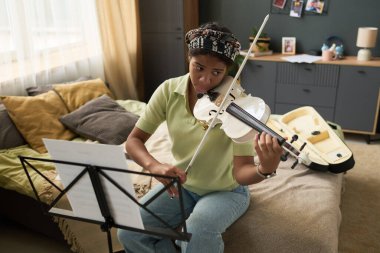 Kiz, yakindaki müzik sehpasinda rahat bir koltukta keman caliyor. Enstrümanlar ve mobilyalar samimi ve kişisel bir hava veriyor.