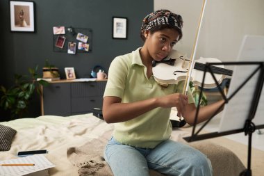 Düşünceli bir ifadeyle rahat bir odada otururken keman çalan kız müzik sehpasındaki notalara odaklanıyor.