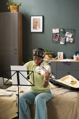 Genç insan yatak odasında keman çalıyor, müzik sehpası ile özenle alıştırma yapıyor daha iyi bir manzara deneyimi için ayarlanmış. Fotoğraf çerçeveleri ve saksı bitkisi atmosferini güçlendiren rahat oda dekorasyonu