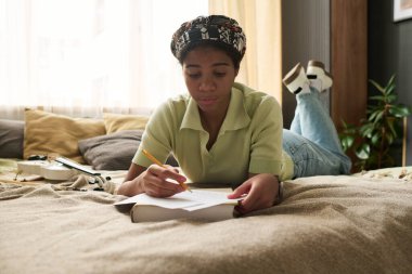 Genç bir kız yatakta uzanıyor ve hoş bir ortamda düşünceli bir ifadeyle notlar yazıyor. Rahatlamaya ve yaratıcılığa odaklan, rahat bir iç mekan ortamı olsun.