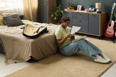 Kız yerde oturuyor, yanında bavul ve gitarla evrak okuyor rahat bir odada bitkiler ve mobilyalar, huzurlu bir atmosfer yaratıyor.