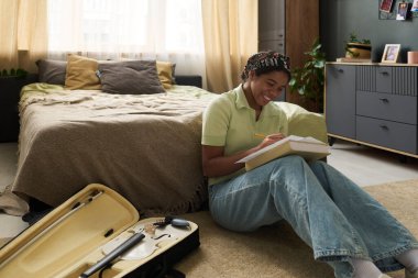 Afro-Amerikalı bir kız deftere yazarken gülümsüyor. Konforlu yatak odasında yerde oturuyor. Etrafı müzik ekipmanları ve yumuşak yataklarla çevrili.