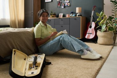 Genç kız yatak odasında gitar kılıfının yanında otururken notlar yazıyor. Çalışma alanı ev bitkileri, fotoğraflar ve biblolarla dekore edilmiş.