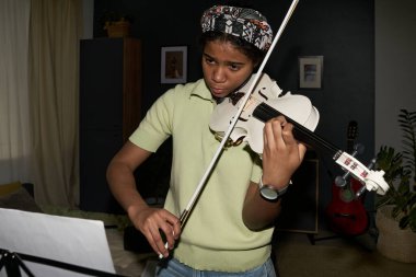 Gençler rahat oturma odasında keman çalmaya odaklanırken müzik sayfaları ayakta tutuluyor. Dekorasyon, yeşil bitkiler ve duvardaki enstrümanlarla sakin bir atmosfer sağlıyor.