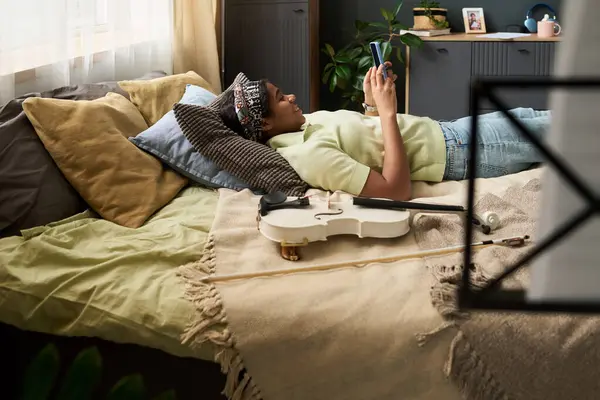 Genç kız yatakta dinleniyor, akıllı telefonları kontrol ediyor. Odada hafif ışıklandırma, yatakta keman ve sakin bir ortam yaratan dekoratif bitkiler var.