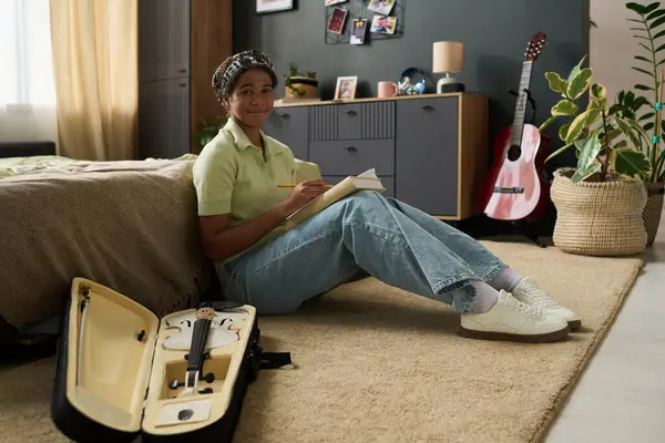 Genç kız yatak odasında gitar kılıfının yanında otururken notlar yazıyor. Çalışma alanı ev bitkileri, fotoğraflar ve biblolarla dekore edilmiş.