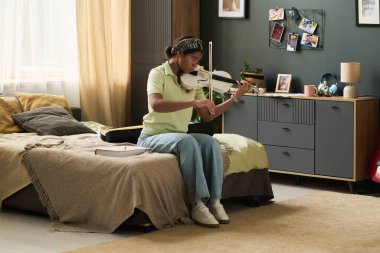 Güzel bir yatak odasındaki battaniyelerle kaplı temiz bir yatakta oturmuş, resim ve resim sergileyen genç bir kız keman çalıyor.