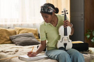 Kız, rahat bir odada, üzerinde çiçekler ve yastıklar olan bir deftere notlar yazarken beyaz keman tutuyor. Doğal güneş ışığı alanı aydınlatıyor.