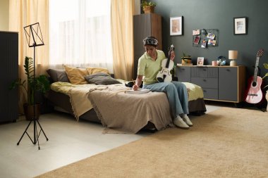 İyi dekore edilmiş yatak odasında gitar çalan kişi doğal ışık, kişisel dekor ve müzik aletleri sergiliyor. Duvarda çeşitli eşyalar bulunan resimler ve raflar var.