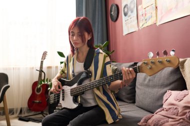 Kızıl saçlı genç kız, duvarında müzik posterleri ve bitkilerin atmosferini yükselten bitkilerle birlikte salonda bas gitar çalıyor. Kızın portresi doğrudan kameraya bakıyor.